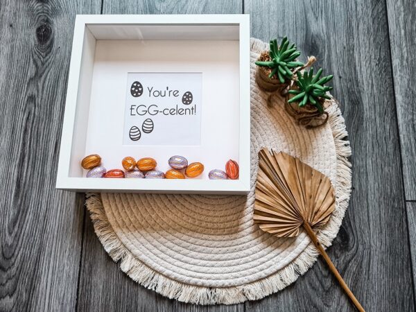 Houten witte fotolijst met de tekst: You're EGG-celent en gevuld met paaseieren