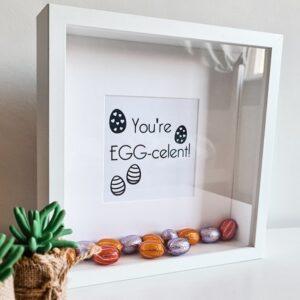 Houten witte fotolijst met de tekst: You're EGG-celent en gevuld met paaseieren